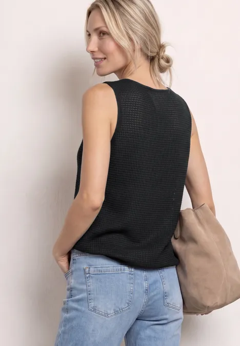 Vrouw van achteren in een zwart mouwloos top, blauwe jeans en een beige schoudertas tegen een lichte achtergrond.