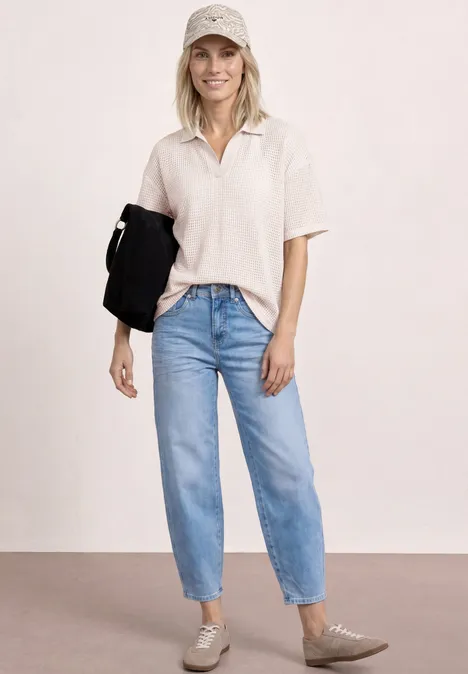 Eine schlanke Frau trägt ein helles Polo, blaue Jeans, beige Schuhe und hält eine schwarze Jacke.