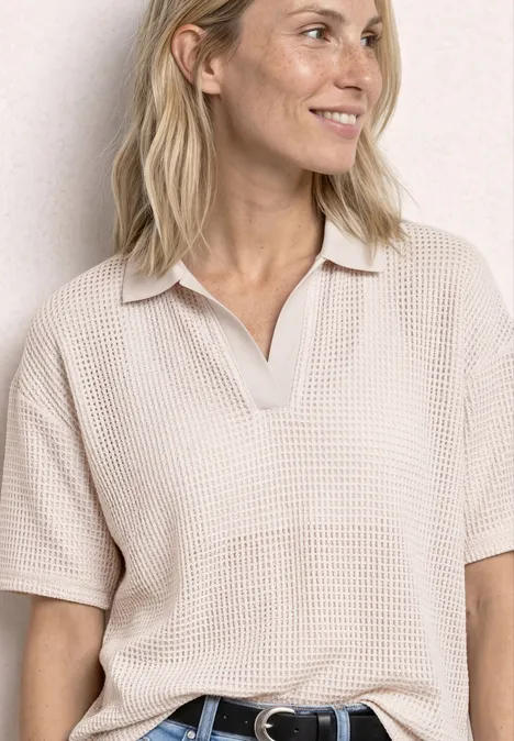 Frau in beigem, kurzärmligem Waffelpolo mit V-Ausschnitt und schwarzem Gürtel vor heller Wand.