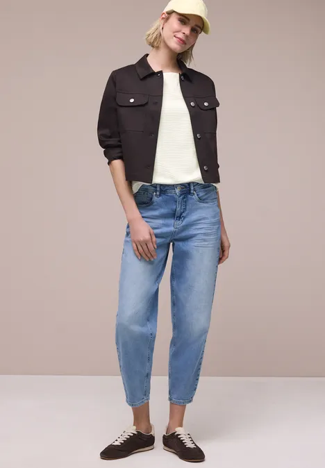 Person in heller Bluse, dunkler Jeansjacke, hellblauen Jeans und braunen Sneakern vor beigem Hintergrund