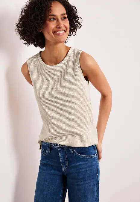 Person steht vor weißem Hintergrund, trägt ärmelloses beiges Oberteil und blaue Jeans.