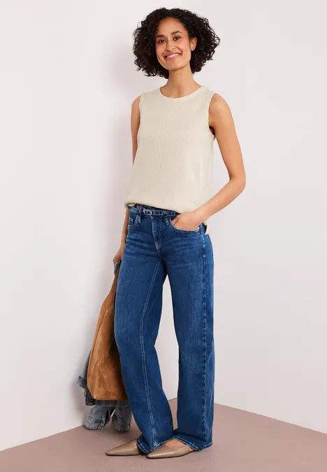 Frau in cremefarbenem Top und weitem Jeans steht lässig vor Wand, Hand in Tasche.