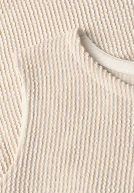 Naheaufnahme eines cremefarbenen, gerippten Strickoberteils mit Rundhalsausschnitt.