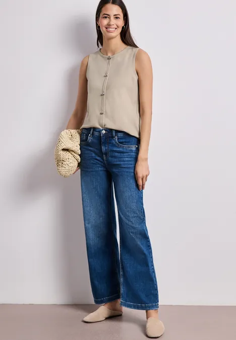 Frau in beige Top und weiten Jeans, mit Strohtasche am Arm und beigen Maultieren.