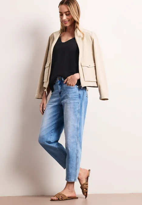 Frau im schwarzen Top, beige Jacke, hellen Jeans und braunen Sandalen vor hellem Hintergrund.
