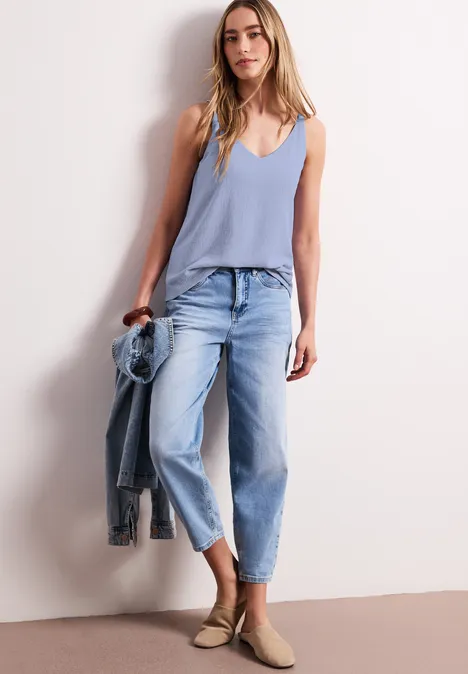 Frau in hellem Top, bequemen Jeans und Espadrilles lehnt an Wand, hält eine Jeansjacke.