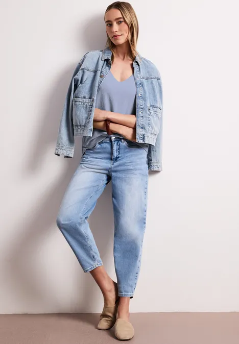 Frau in hellem Denim-Outfit: V-Neck, Jeans, Jacke locker über die Schultern gelegt, beige Slipper.