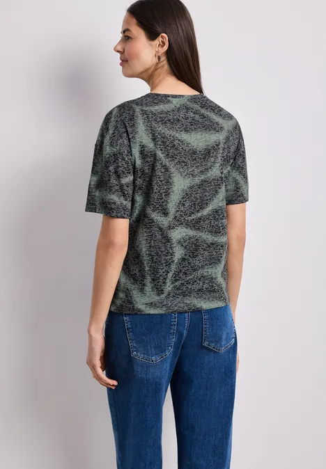 Eine Frau steht mit dem Rücken zur Kamera und trägt ein grün-schwarzes Muster-T-Shirt mit blauer Jeans.