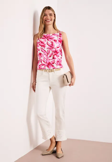 Vrouw in een roze-wit bloemen-top, witte ¾-broek met borduursel, beige pumps en een clutch in de hand.