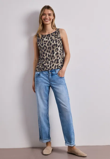 Frau steht vor weißer Wand, trägt ärmelloses Top mit Leopardenmuster, blaue Jeans und beigen Schuhen.