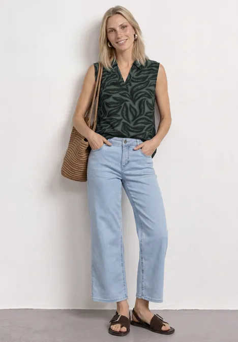Top mit Kragen und Strukturmix deep bound green Frau in ärmellosem grünen Blattmuster-Top, hellblauen, hochgeschnittenen Jeans und braunen Sandalen mit Strohtasche.