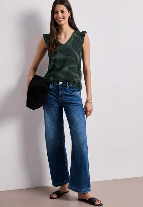 Frau vor weißer Wand in ärmellosem grünen Top, weiten Jeans, schwarzen Sandalen und mit schwarzer Tasche.