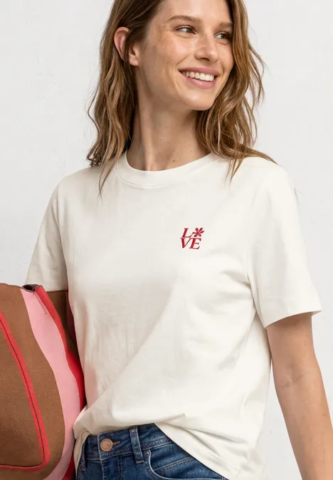 Frau trägt ein weißes T-Shirt mit kleinem 'LOVE'-Schriftzug, Jeans und einer rosa-braunen Tasche.