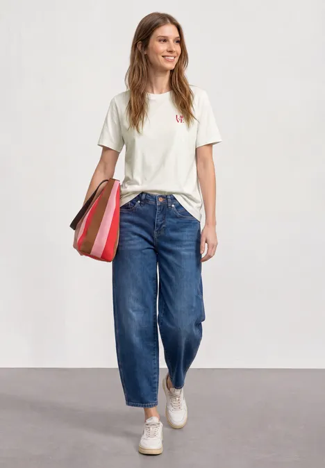 Frau trägt weißes T-Shirt, blaue Jeans und weiße Sneaker, hält gestreifte Tasche vor hellem Hintergrund.