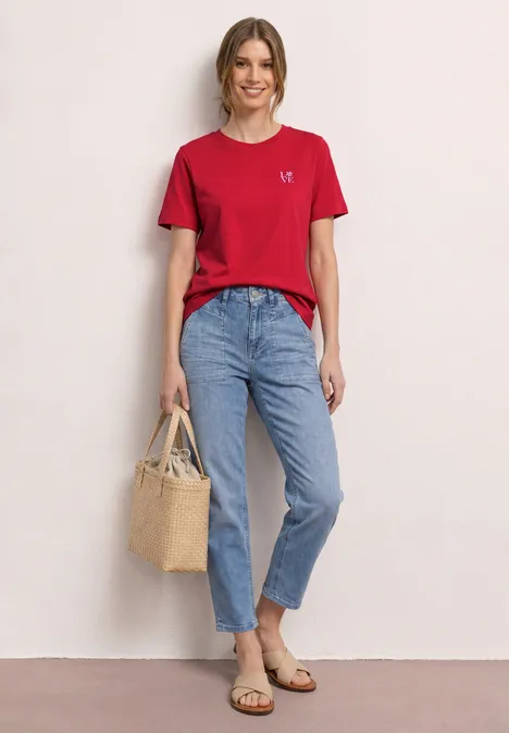 Frau posiert vor neutralem Hintergrund in rotem T-Shirt, lockeren Jeans, flachen Sandalen und Korbtasche