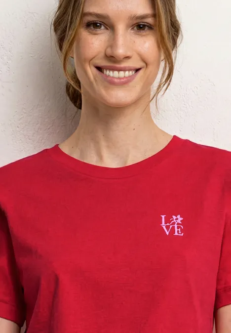 Person in rotem T-Shirt mit kleinem weißen 'LOVE'-Aufdruck vor weißer Wand.