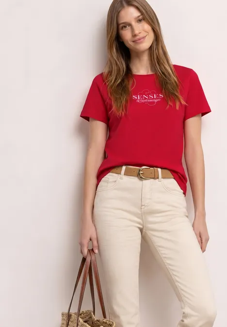 Een vrouw in een rood T-shirt, beige broek met ceintuur, die een handtas vasthoudt.