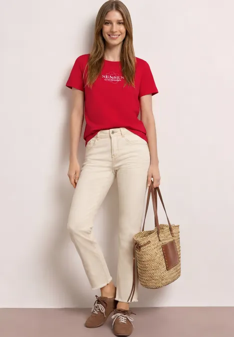 Vrouw in rood T-shirt, beige broek en bruine schoenen, met een rieten tas voor een neutrale achtergrond.