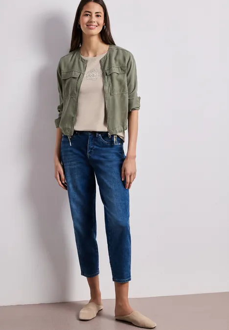 Frau trägt khakigrüne Hemdbluse, beiges Top, blaue Jeans und beigen Ballerinas vor hellem Hintergrund.