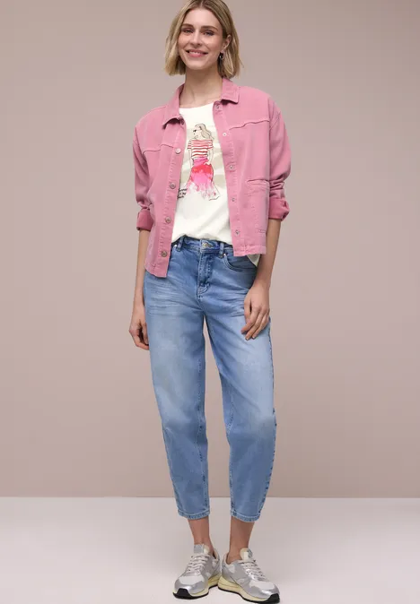 Modell trägt pinke Jeansjacke, bedrucktes weißes T-Shirt, lockere blaue Jeans und Sneaker vor neutralem Hintergrund.
