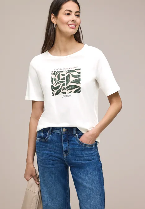 Een vrouw draagt een wit T-shirt met groen dessin en een blauwe spijkerbroek.