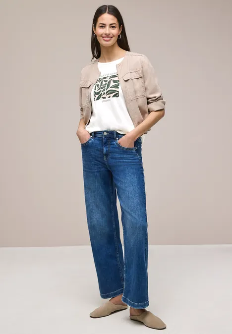 Vrouw voor neutrale achtergrond met licht jasje, wit T-shirt met groene print, wijde jeans en platte schoenen.