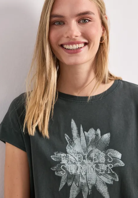 Frau mit langen blonden Haaren in schwarzem T-Shirt mit verblasstem Sonnenblumen-Motiv und Schriftzug vor weißem Hintergrund