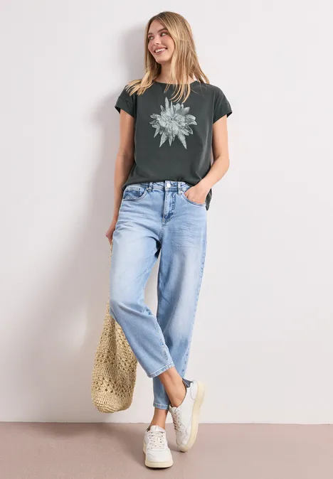 Frau trägt graues T-Shirt mit Blumenprint, helle Jeans, Sneakers und Strohtasche, steht entspannt.