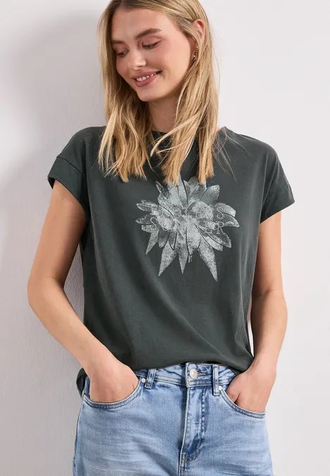 Frau in dunklem T-Shirt mit floralem Blattmotiv und blauen Jeans, Hände in den Hosentaschen.