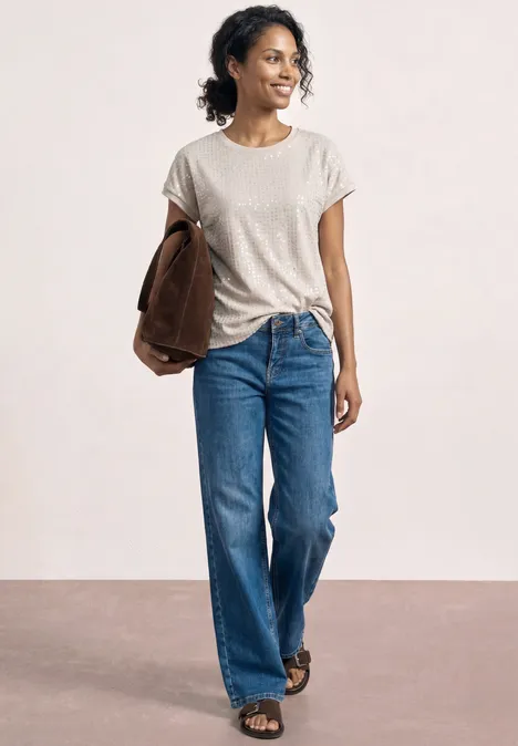 Person trägt weißes T-Shirt, blaue Jeans, Sandalen, hält eine braune Jacke lässig unterm Arm.