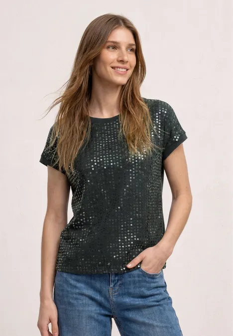 Eine Frau trägt ein grünes Paillettent-Shirt mit kurzen Ärmeln und blaue Jeans, steht vor hellem Hintergrund.