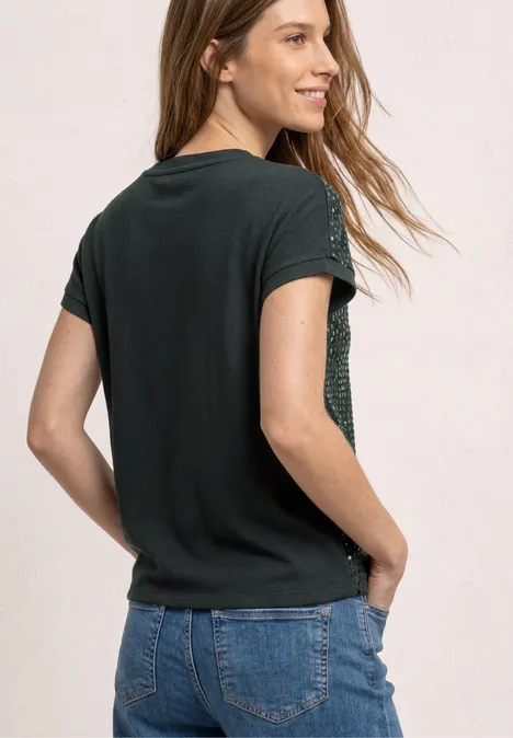 Junge Frau mit schwarzem T-Shirt und Jeans steht seitlich, Hände in den Taschen, vor hellem Hintergrund.