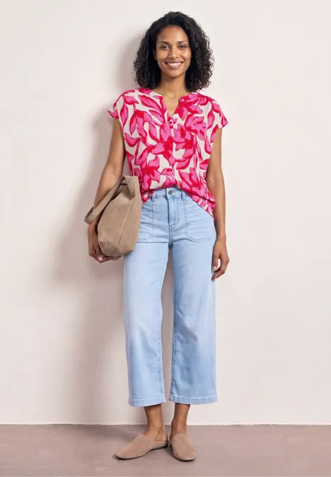 Frau in pink gemusterter Bluse, hellblauen Culotte-Jeans, beigem Shopper und Loafers.