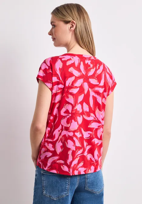Frau von hinten in roter Bluse mit rosa Blattmuster und blauer Jeans.