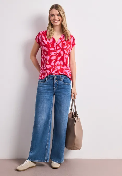 Frau in rotem Muster-Shirt, weiten Jeans, weißen Schuhen und brauner Tasche vor hellem Hintergrund.
