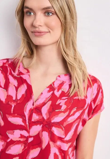 Frau trägt ein rosa-rotes, blumiges Oberteil vor weißem Hintergrund.
