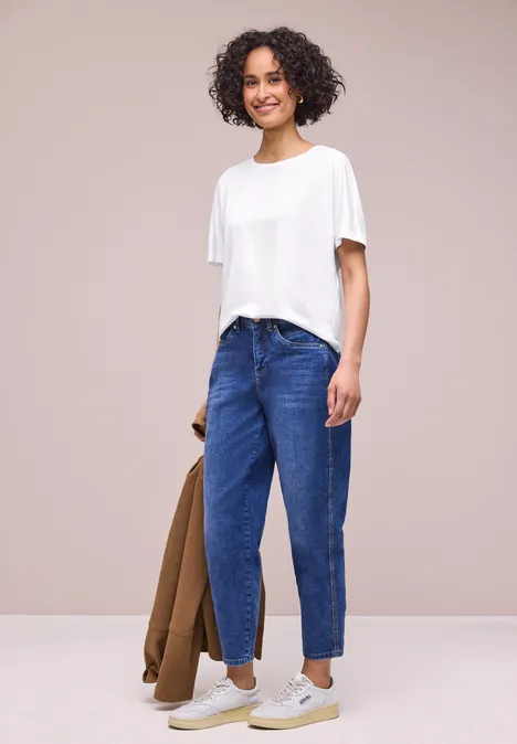 Person trägt weißes T-Shirt, blaue Jeans und hält eine braune Jacke vor hellem Hintergrund.