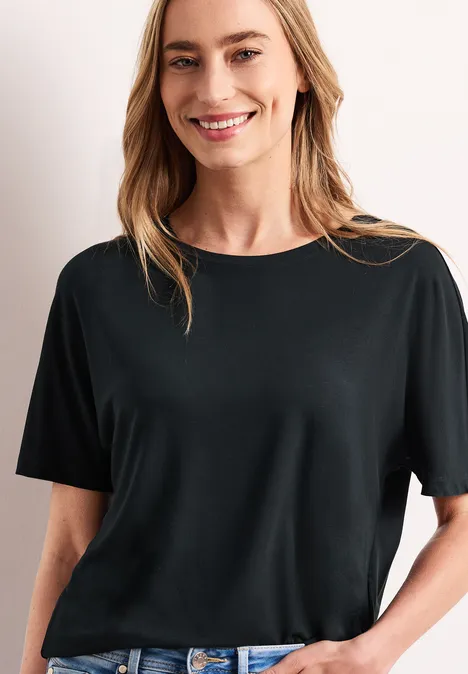 Eine lächelnde Frau in schwarzem T-Shirt und Jeans, steht lässig vor hellem Hintergrund.