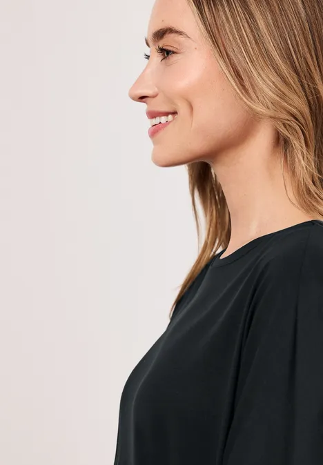 Profilaufnahme einer lächelnden Frau mit blonden Haaren und schwarzem Oberteil vor hellem Hintergrund.