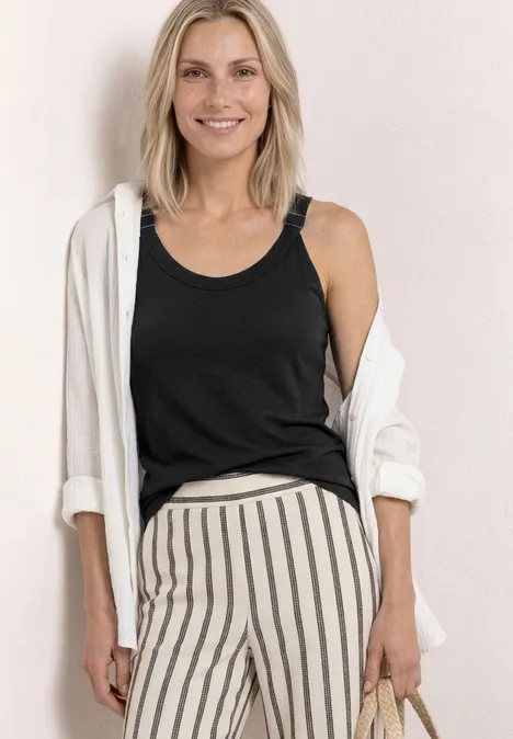 Frau in schwarzem Tanktop, weißer Hemdjacke und gestreifter Hose mit Tasche in der Hand.