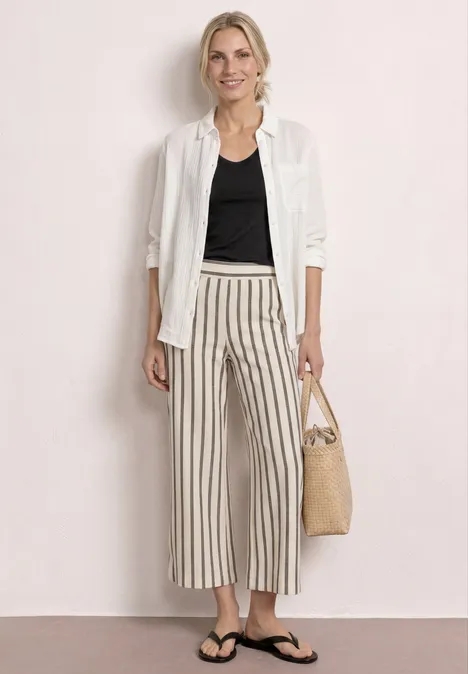 Frau mit weißem Hemd über schwarzem Top, gestreiften Culotte-Hosen, Flip-Flops und Korbtasche vor neutralem Hintergrund.