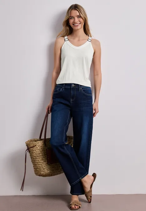 Eine Frau trägt ein weißes Top, dunkle Jeans, Espadrilles und einen Bastkorb vor hellem Hintergrund.