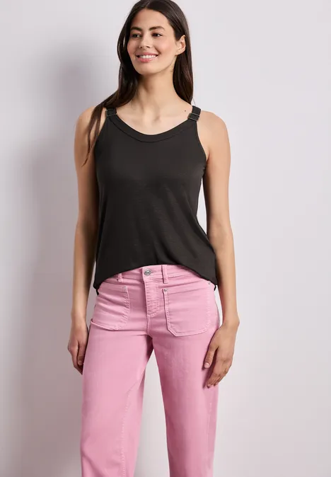 Eine Frau in schwarzem Tanktop und pinken Jeans steht vor neutralem Hintergrund.