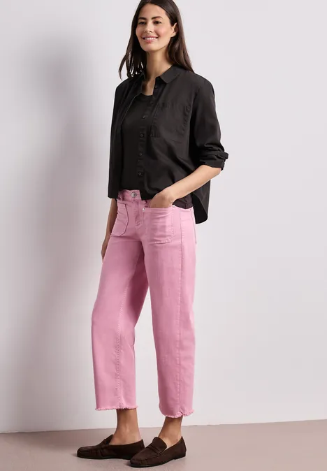 Eine Person trägt eine schwarze Bluse und rosa gerade geschnittene Hosen vor schlichtem Hintergrund.