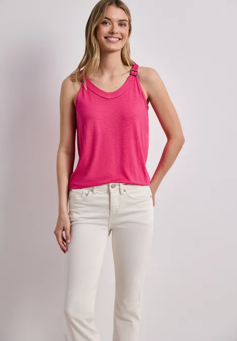 Frau trägt pinkes Tanktop und weiße Jeans, steht vor neutralem Hintergrund.