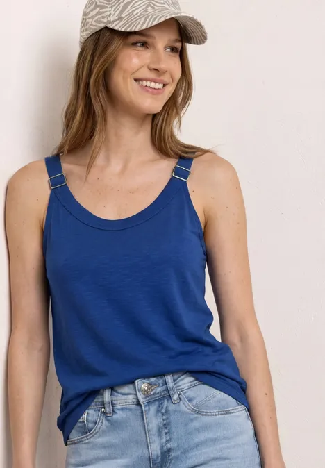Frau mit blauem Top, Jeans und Cap vor neutralem Hintergrund.