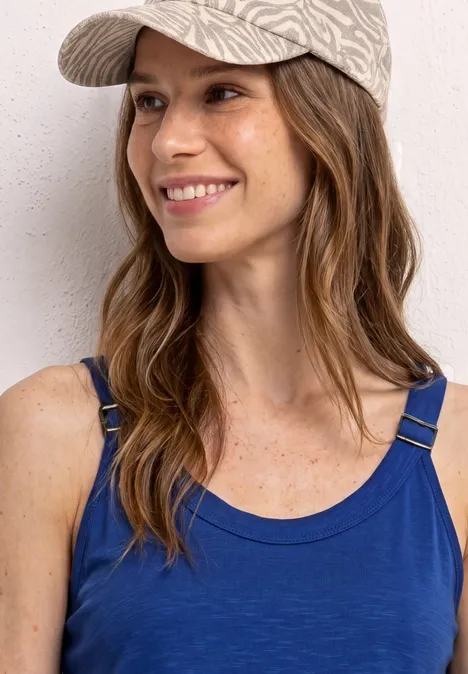 Eine Person trägt ein blaues Trägershirt und eine gemusterte Kappe vor grauem Hintergrund.