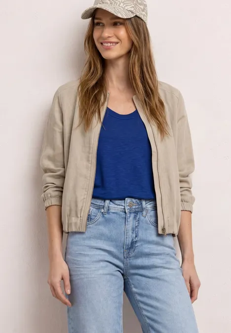 Frau in beiger Jacke, blauem Top und Jeans, stehend vor heller Wand.