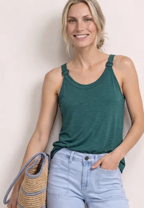 Person mit grünem ärmellosem Top und hellblauer Jeans hält eine geflochtene Tasche aus Stroh.