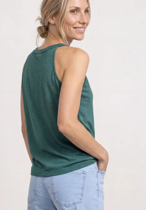 Eine Frau in grünem ärmellosem Top und hellblauer Jeans, lächelnd mit den Händen in den Taschen.
